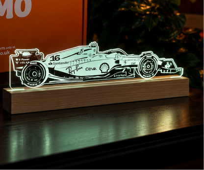 A LUMO LAMP brand LED light shaped like a Formula 1 car, glowing on a wooden base in a dark indoor setting, with its orange branded box behind it.